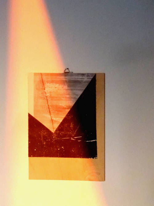 A laser print paper collage on wooden board. It shows a geometric composition with different grayscale printed areas. Parts of the wooden board are visible, as the paper doesn’t fill the whole board. The picture was taken with the work on tha wall, illuminated by rays of the setting sun.